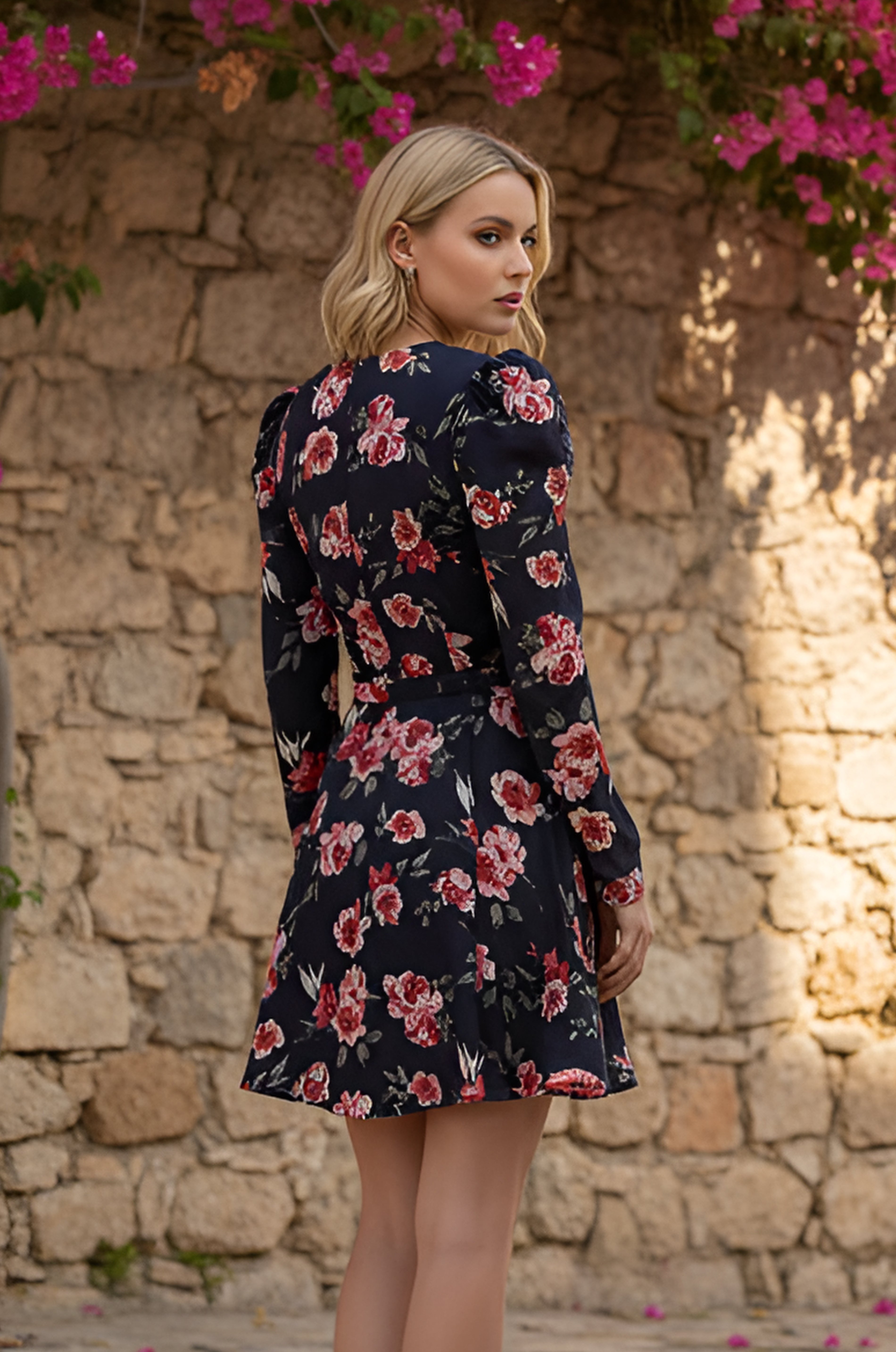 A woman with blonde hair wears the Midnight Rose Sleeved Short Casual Dress: Harper, standing outdoors before a stone wall with pink flowers blooming above.